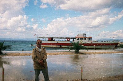 Vintage seaside scene featuring a man in a beige trench coat by a shallow, reflective waterfront. A red-and-white ferryboat w...