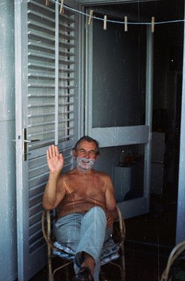 Mid-century bathroom interior with closed louvered shutters, featuring a man seated on a wicker chair, shirtless with lathere...