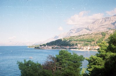 Vintage coastal scene featuring a serene bay framed by lush greenery and rugged mountains. A small coastal village with clust...