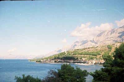 Vintage coastal scene with steep forested mountains framing a small seaside village. Dense greenery meets turquoise waters un...