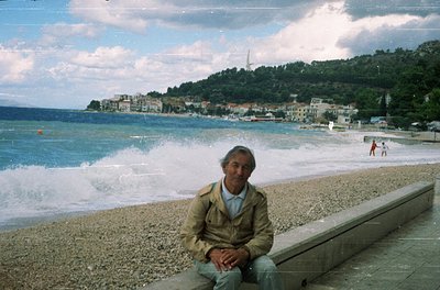 Man in vintage jacket sits on concrete ledge by seaside, overlooking coastal town with waves crashing. Mid-20th century beach...