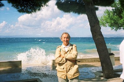 Man in light beige jacket poses by seaside with ocean waves crashing in background. Mid-20th century coastal setting, likely ...
