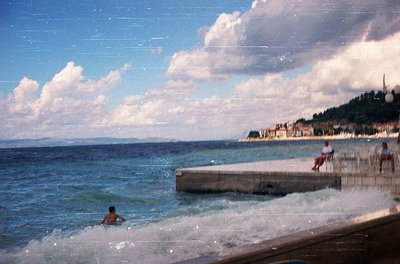 Vintage seaside scene with a man swimming in shallow waves near a concrete pier. Two figures sit on the edge, overlooking the...