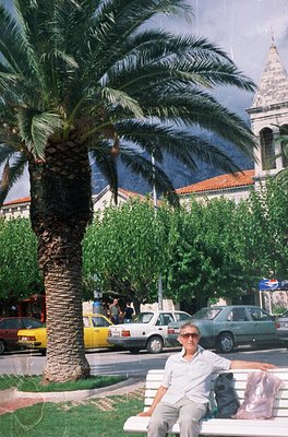 A man sits on a concrete bench beside a palm tree in a Mediterranean-style plaza, overlooking a historic building with arched...
