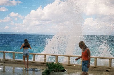 Two women enjoy a seaside fountain’s spray on a sunny day, likely mid-20th century. One wears a red tank top and denim shorts...