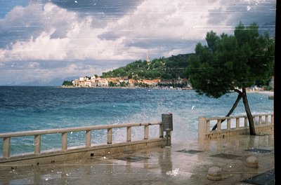 Vintage coastal scene featuring a seaside promenade with wooden railings and wet pavement. Distant hillside town with Mediter...