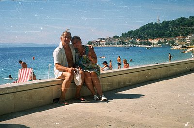 Couple posing on a concrete pier by a seaside resort, mid-20th century. Man in light linen shirt, woman in floral dress. Back...