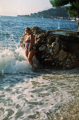 Vintage seaside moment: woman in floral one-piece swimsuit steps into shallow waves near rocky shore. Coastal greenery and di...