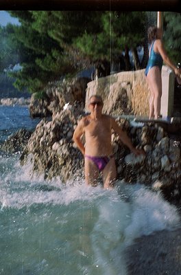 A man in purple swim trunks stands in a natural rock waterfall pool by a seaside cliff, with water cascading around him. Anot...
