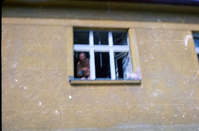 A close-up of a worn, yellowish plaster wall with a small, square window featuring six panes. Inside, a person’s face is part...