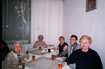 Five adults seated indoors at a round table, enjoying drinks and conversation. The setting appears to be a modest, well-lit r...