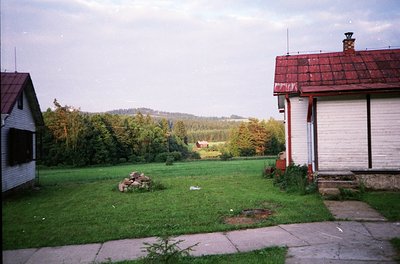 Mid-century suburban landscape featuring two single-story houses with red-tiled roofs and white walls. Overgrown grass and a ...
