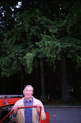 Mid-20th century man in a forest setting, holding a red pushcart. He wears a light-colored jacket, dark trousers, and a white...