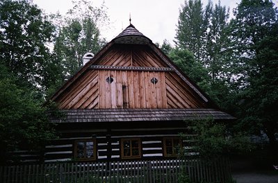 Traditional wooden house with steep, gabled roof and log construction, set in a forested area. Dark brown logs with white win...