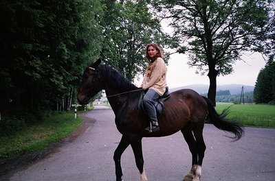 Woman riding a dark brown horse along a tree-lined road, dressed in a beige jacket and jeans. Lush greenery and misty hills i...
