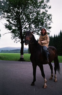 A woman in casual 1990s attire rides a dark-coated horse on paved road, flanked by lush greenery and distant forested hills. ...