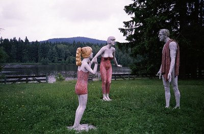 Three life-sized plaster sculptures in outdoor setting, likely 1960s–70s. Female figure in red knit dress extends hand toward...