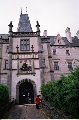 Neogothic-style entrance gate with stone carvings of a knight on horseback and heraldic symbols. Two people in red jackets st...