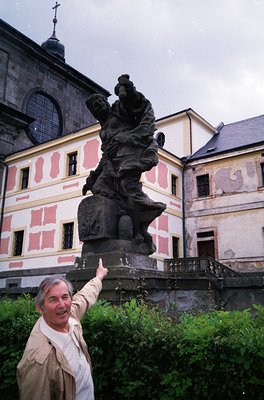 A man poses beside a dramatic bronze statue of a robed figure with outstretched arms, perched atop a pedestal in a courtyard....