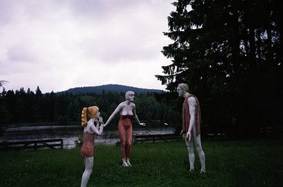 Vintage sculpture installation featuring three human-like figures in outdoor setting. Left figure wears a red dress, center f...