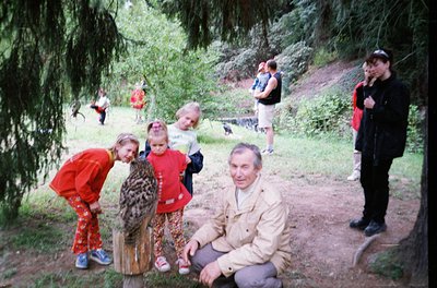 Vintage outdoor group photo in a wooded area, featuring a man in a beige jacket squatting with two young girls in patterned c...