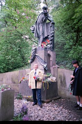 Monumental bronze statue of a robed figure in a park setting, surrounded by floral tributes. A man in a beige coat holds a bo...