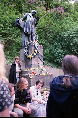 Bronze statue of seated figure in classical attire, surrounded by floral wreaths and bouquets at a memorial site. Group of pe...