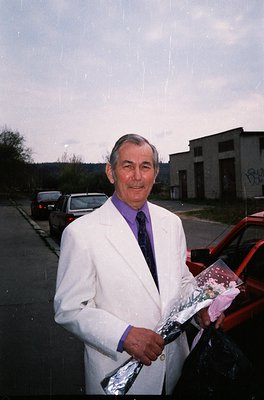 Mid-20th century man in formal attire (white suit, purple tie) holding a wrapped gift, likely a celebratory occasion. Urban i...