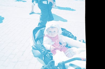 Vintage sepia-toned photo of an infant in a classic stroller on a paved surface, likely mid-20th century. Child wears a white...
