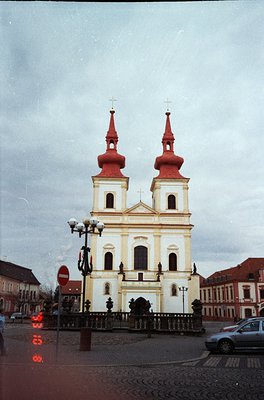 Baroque-style church with twin red-domed towers, cream façade, and symmetrical windows—likely Eastern European. Cobblestone p...