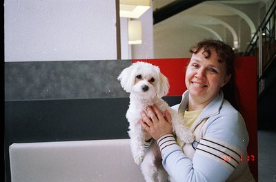 Vintage candid of a woman holding a small white poodle in an indoor setting, likely a public transport hub. She wears a strip...