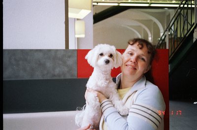 Vintage indoor portrait of a woman holding a small white poodle-type dog, likely 1980s-1990s. Staircase and geometric wall pa...
