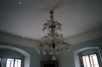 Vintage chandelier with ornate crystal branches suspended in a dimly lit, classical-style room. Symmetrical arched windows wi...