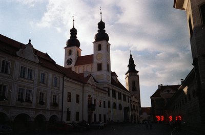 Baroque-style church with twin towers and clock, flanked by arched buildings in a European plaza. Red digital sign reads "199...