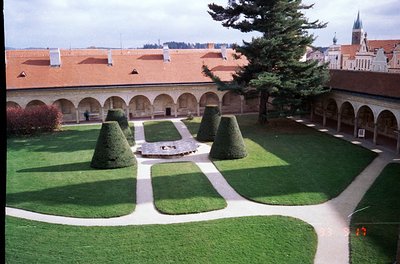 Neoclassical cloister courtyard with symmetrical topiary gardens, gravel pathways, and arched colonnades. Red-tiled roofs and...