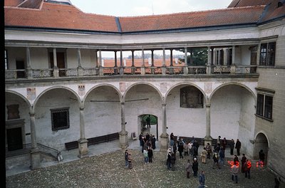 Two-story courtyard with arched colonnades, red-tiled roof, and weathered plaster walls. Group of people gathered near centra...