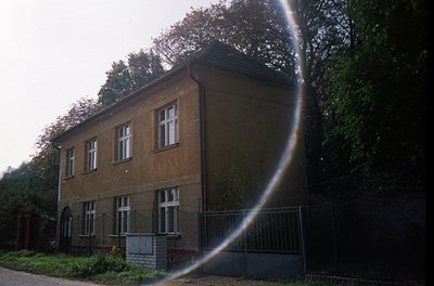 Two-story brick building with symmetrical windows, surrounded by chain-link fence. Lens flare obscures part of the frame. Lik...