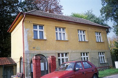 Two-story yellow-brick residential building with white-framed windows, showing signs of aging. Red car parked near a gated en...