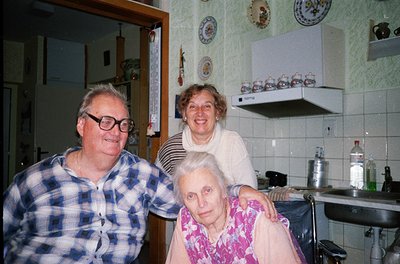 Mid-20th century kitchen interior with three elderly individuals posing. Man in checkered shirt, woman in striped sweater, se...
