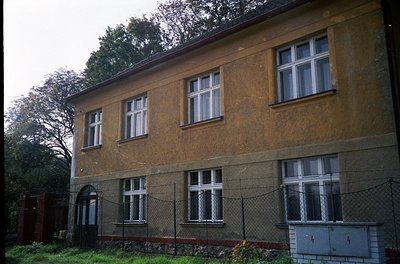 Two-story brick building with aged yellowish facade, featuring symmetrical white-framed windows. Chain-link fence with red br...