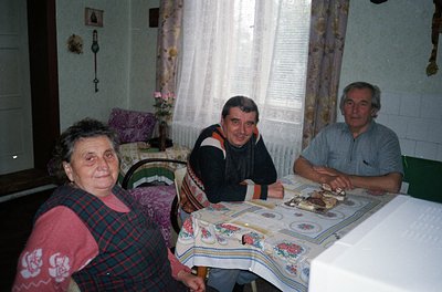 Three adults seated indoors at a table covered with a floral-patterned tablecloth, likely in a rural or traditional home sett...