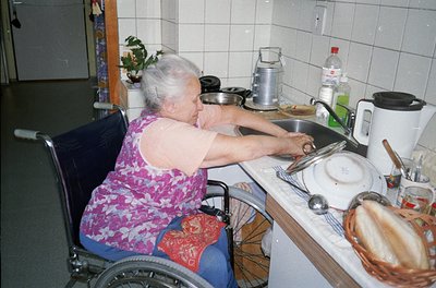 Indoor kitchen scene featuring an elderly woman in a wheelchair preparing food. She wears a floral-patterned sleeveless top a...