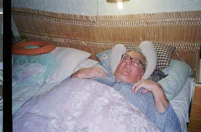 A relaxed elderly man reclines on a floral-patterned bed with a white bunny pillow propped behind his head. The headboard fea...