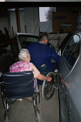 Couple assisting elderly woman into a car via wheelchair at a gas station with "Tango" branding. Indoor/outdoor lighting sugg...