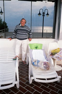Man in striped sweater and light trousers poses on a seaside balcony overlooking waves, seated on plastic lounge chairs with ...