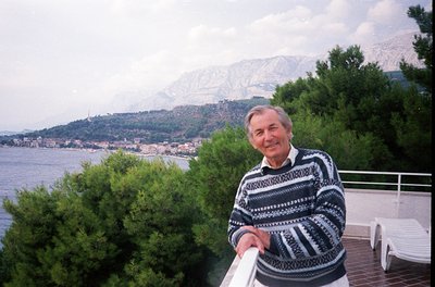 Man in a patterned sweater poses on a seaside balcony overlooking a coastal town and mountainous backdrop. Mid-20th century M...