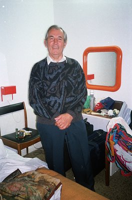 Mid-century hotel room interior featuring a man in a patterned sweater and dark trousers. Minimalist furnishings include a sm...