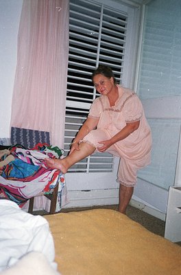A woman in a light pink housecoat assists a seated individual with a leg elevation exercise, likely post-surgery or injury. T...