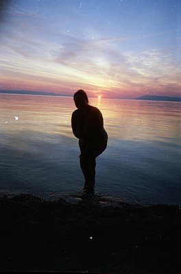 Silhouetted figure stands at water’s edge during golden-hour sunset, back to camera. Warm tones reflect on calm waters, likel...