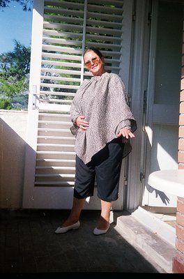 Woman in a patterned, shawl-collared blouse with cropped, wide-leg trousers and white sneakers, posing on a sunlit porch with...